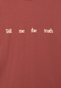 T-shirt de couleur rouille avec du texte blanc en trois dimensions épelant "Dis-moi la vérité" sur la poitrine dans une police ludique et courbée.