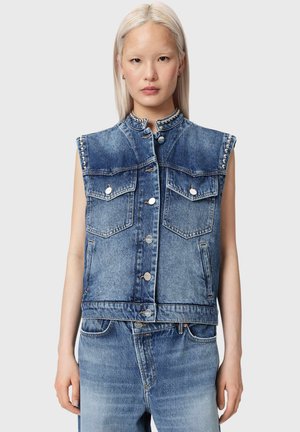 Młoda kobieta z prostymi blond włosami, ubrana w bezrękawnik z dżinsu z ćwiekami i pasujące wyblakłe niebieskie dżinsy, stojąca na jednolitym tle.