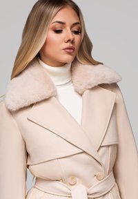 Mladá žena s dlouhými blond vlasy má na sobě béžový kabát s širokým huňatým límcem a bílý rolák.