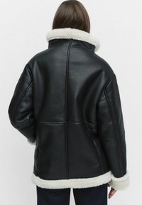 Svart läderjacka med en tjock vit pälskrage och manschetter. Har en fyrkantig design och synlig sömdetalj på baksidan.