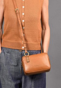 Bolso cruzado de cuero marrón con forma rectangular, correa ajustable, detalles en hardware dorado y cierre de cremallera.
