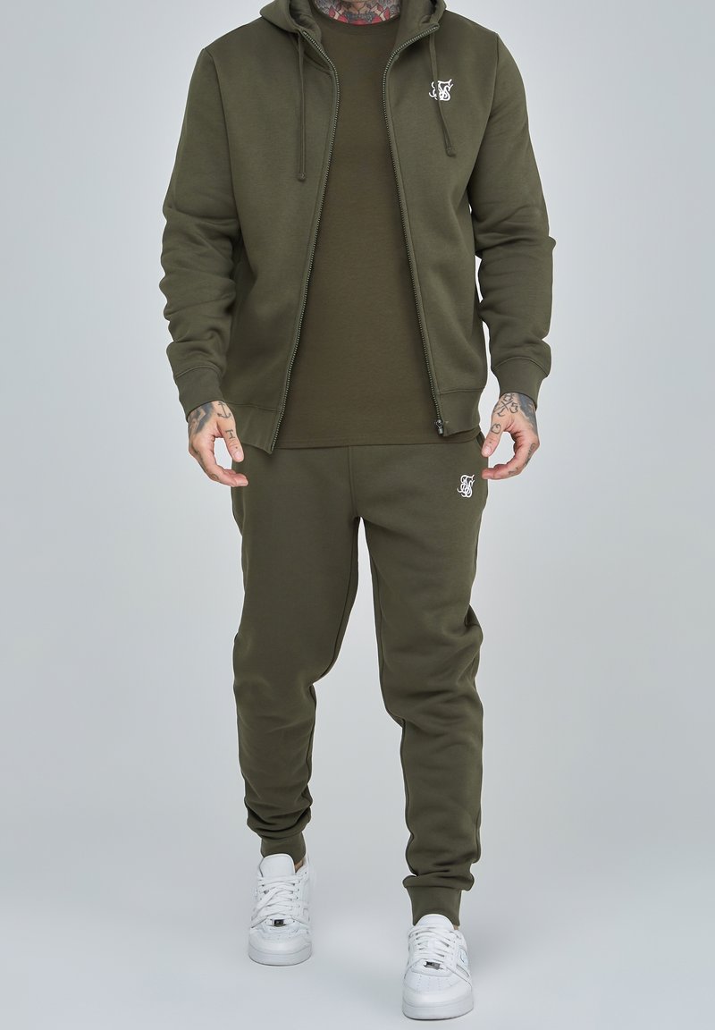 Sudadera de color verde oliva con cremallera, joggers a juego en verde oliva, detalle del logo en blanco en el pecho y la pierna, y zapatillas blancas. Tejido de mezcla de algodón.