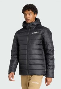 Casaco acolchoado preto com capuz, com design matelassê, fecho de correr frontal e logótipo no peito esquerdo. Textura suave, isolamento térmico.