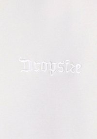 Weißer Stoff mit gesticktem Text "Dropsize" in einer gotischen Schriftart, mit einer glatten, matten Textur und ohne zusätzliche Muster oder Akzente.