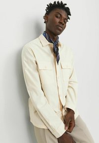 Chemise boutonnée de couleur claire avec deux poches poitrine, manches longues et coupe décontractée, accompagnée d'une écharpe bleue à motif autour du cou.