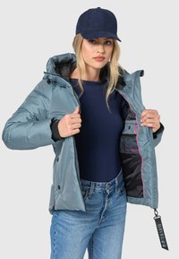 Lichtblauwe pufferjack met een capuchon, zwarte voering en roze accenten. Geopend om de binnenzak en het logo-tag te tonen. Gecombineerd met een marineblauw T-shirt en jeans.
