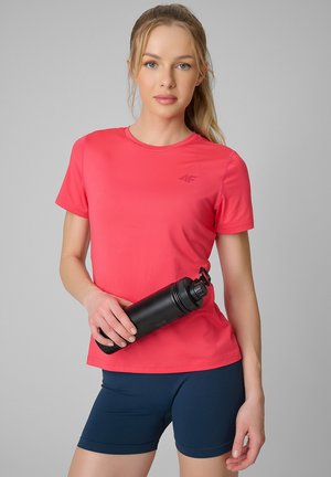 Kobieta z blond włosami związanymi w kucyk, ubrana w koralowy t-shirt z krótkim rękawem i granatowe szorty, trzyma czarną butelkę na wodę na szarym tle.