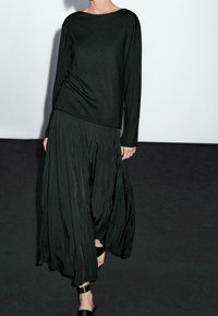 Musta neulottu pitkähihaisen yläosa, jossa on leveä pääntie, yhdistettynä laskeutuvaan, laskostettuun mustaan hameeseen. Molemmilla vaatekappaleilla on teksturoitu pinta.