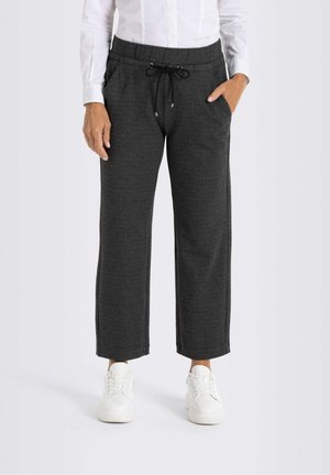 Henkilö, jolla on tummanharmaat kapealahkeiset housut, joissa on kiristysnauha vyötäröllä, valkoinen napillinen paita ja valkoiset lenkkarit, seisoo käsi taskussa.