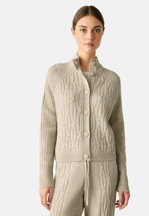 Frau trägt eine beige kabelgestrickte, geknöpfte Strickjacke und passende gestrickte Hose, steht vor einem schlichten hellen Hintergrund.