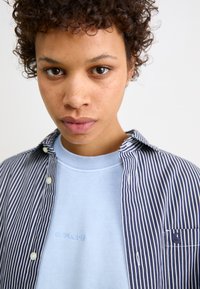 Chemise à rayures bleu marine et blanche sur un sweat-shirt bleu clair avec logo brodé. Le modèle a les cheveux bouclés et un fond neutre.