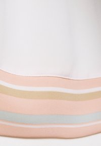 Vit topp i tyg med en jämn textur; fållen har horisontella ränder i ljusrosa, mjuk blå och guldaccenter.