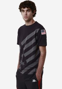 T-shirt nera con strisce diagonali grigie e stelle, con una piccola patch della bandiera americana sulla manica sinistra. Tessuto in cotone morbido.
