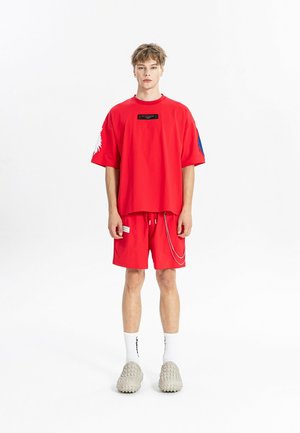 Joven de pie, con camiseta roja brillante de gran tamaño y pantalones cortos a juego con detalle de cadena, calcetines blancos y zapatos beige texturizados sin cordones.