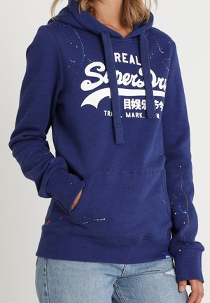 Granatowa bluza z kapturem z białym logo „Superdry”, sznurkami oraz przednią kieszenią, ozdobiona plamami farby, noszona z jasnoniebieskimi dżinsami.