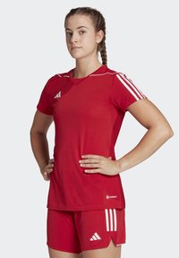 Maglia sportiva rossa con pantaloni corti, entrambi con strisce bianche; la maglia ha un colletto a V e dettagli in rete sulle spalle; design aderente per lo sport.