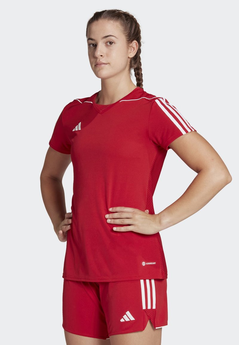 Maglia sportiva rossa con pantaloni corti, entrambi con strisce bianche; la maglia ha un colletto a V e dettagli in rete sulle spalle; design aderente per lo sport.