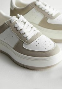 Zapatillas blancas y beige con un upper suave, detalles perforados, cordones planos y una suela gruesa y texturada.