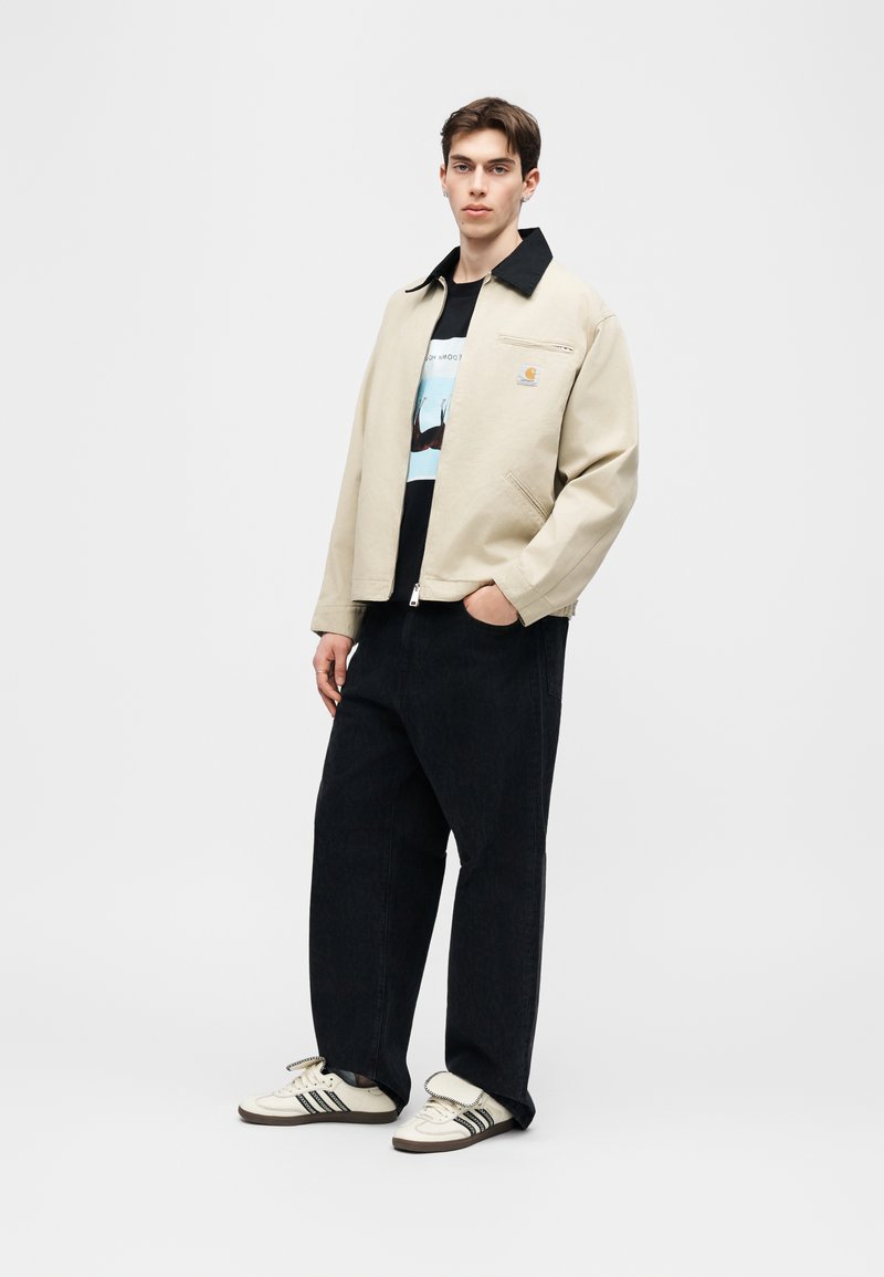 Joven de pie, lleva una chaqueta beige con cuello negro, camiseta gráfica negra, pantalones anchos negros y zapatillas blancas con rayas negras.