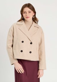Veste en laine mélangée beige à coupe courte avec un design à double boutonnage, des boutons noirs, des revers larges et des poches latérales, associée à un col roulé à côtes.