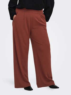 Personne portant un pantalon taille haute, à jambes larges, couleur rouille, les mains dans les poches et des chaussures noires pointues, sur un fond clair uni.