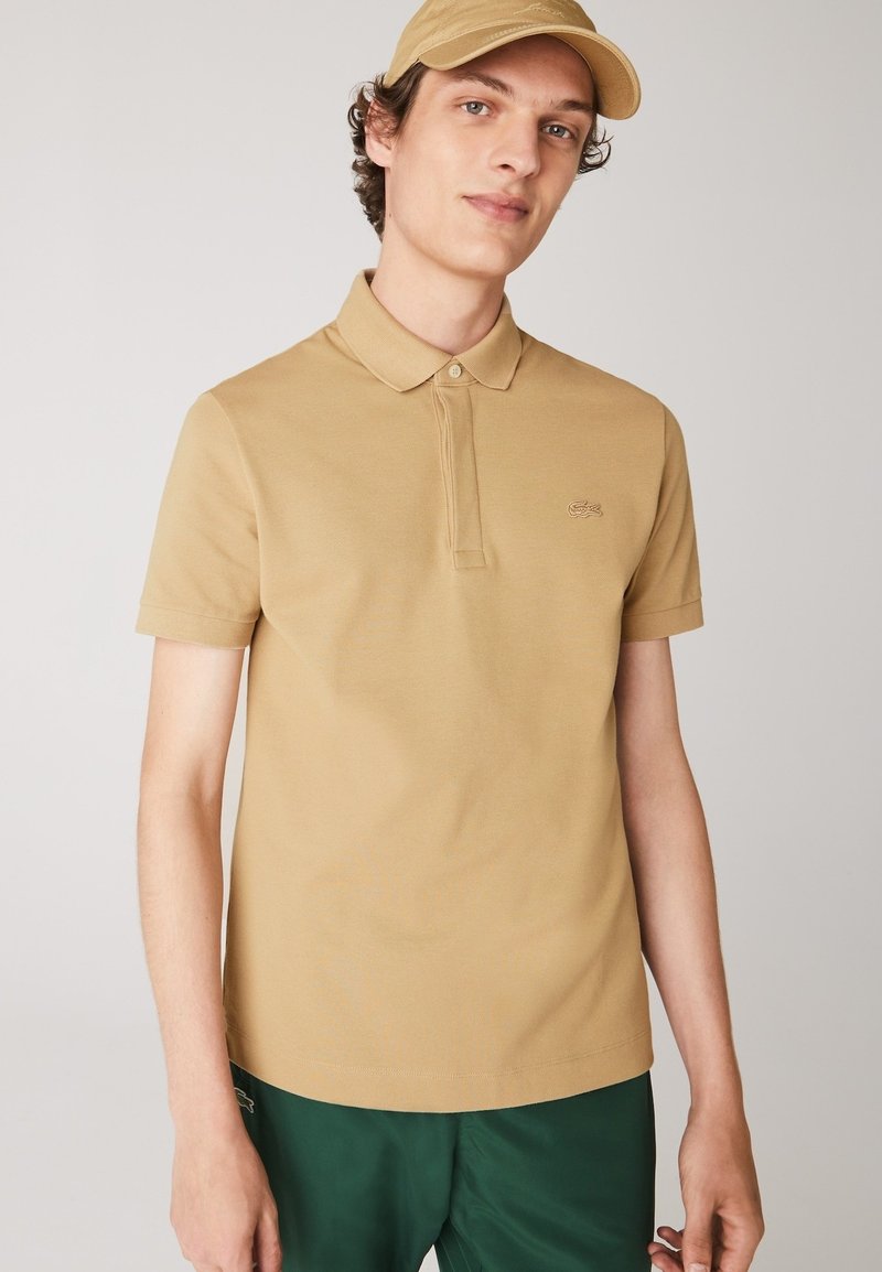 Beige polo skjorta med krage, korta ärmar och en diskret logotyp på vänster bröst. Slät textur, tillsammans med gröna shorts.
