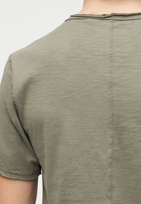 Vue de dos d'une personne portant une chemise à manches courtes de couleur vert olive clair avec des coutures visibles au centre et autour du col.