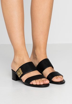 Mules en cuir noir à talon bloc avec deux larges lanières sur le pied, comportant un accent en métal doré avec un détail de logo. Fini lisse et mat.