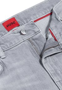 Jeans de denim gris claro con cremallera plateada parcialmente abierta y botón, que presentan una etiqueta interior en la cinturilla roja con el texto "HUGO".