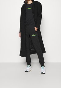 Abrigo largo negro, camiseta negra ajustada con una etiqueta verde, joggers negros a juego, y zapatillas blancas con acentos coloridos.