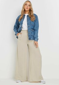 Denim jacket in blue with button front and two pockets, paired with beige wide-leg pants and white sneakers, on a neutral background.