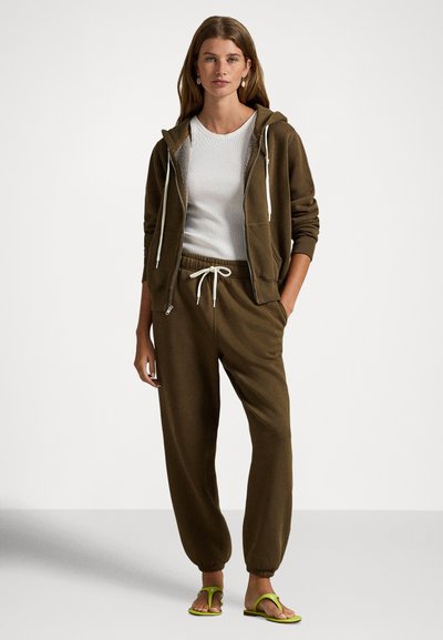 Brun hettejakke med glidelås og snor, hvit ribbestrikket skjorte, matchende brune joggebukser og grønne flip-flops. Glatt stoff, avslappet passform.