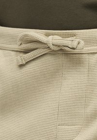 Κοντινό πλάνο μπεζ υφασμάτινου παντελονιού με υφή, με δεμένη κορδόνι στη μέση και μερική τσέπη ορατή στη δεξιά πλευρά.