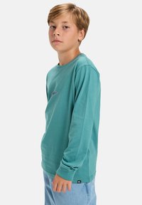Langärmliges türkisfarbenes Hemd aus weicher Baumwolle mit einem minimalistischen grauen Logo und kontrastierenden Nähten. Kombiniert mit hellblauen Jeans.