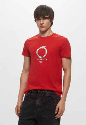 T-shirt in cotone rosso con un logo e un design testuale argentati al centro. Maniche corte e scollo rotondo. Indossato con jeans scuri.