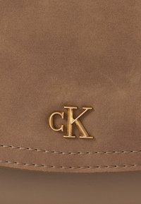 Beżowy zamsz o gładkiej fakturze, z akcentem w postaci złotego logotypu "CK". Detal przeszycia wzdłuż krawędzi nadaje wysublimowane wykończenie.