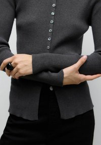 Šedý žebrovaný cardigan s knoflíkovým zapínáním, dlouhými rukávy a mírně zkráceným lemem. Černý prsten na ruce držící opačnou ruku.
