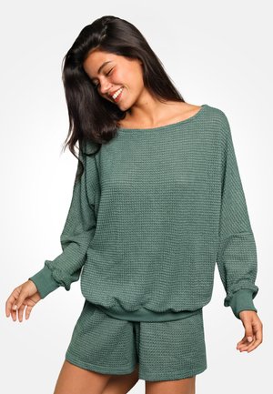 Frau lächelt mit geschlossenen Augen, trägt einen strukturierten grünen Pullover mit langen Ärmeln und passende Shorts vor weißem Hintergrund.
