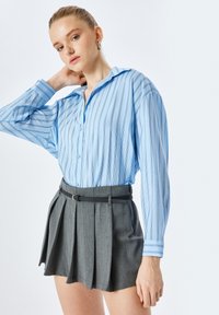 Lichtblauw, verticaal gestreept shirt met een kraag, lange mouwen, en grijze geplooide shorts met een zwarte riem. Eenvoudig ontwerp en maatwerk pasvorm.