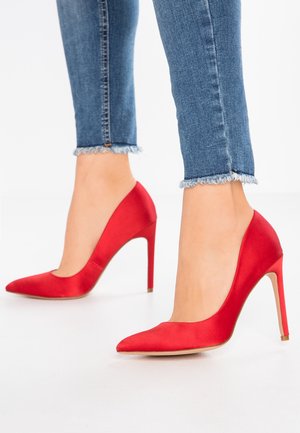 Røde satin højhælede sko med spids tå og stilettohæle. Skoene er parret med denimjeans med frynset kant.
