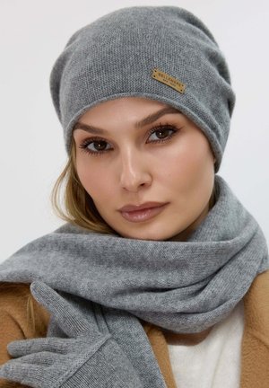Femme portant un bonnet, une écharpe et des gants assortis en maille grise avec un manteau camel sur fond blanc.