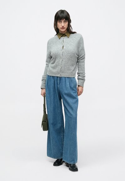 Γκρι πλεκτή ζακέτα πάνω από πουκάμισο σε ελιά πράσινο χρώμα, συνδυασμένη με μπλε τζιν παντελόνια με φαρδιά μπατζάκια και μαύρες μποτάκια αστραγάλου. Περιλαμβάνεται πράσινη τσάντα.