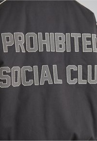 Jacheta neagră cu litere mari alb-negru, texturate cu plasă, care citește „SOCIAL CLUB PROHIBIT” pe spate. Material neted cu un finisaj mat.