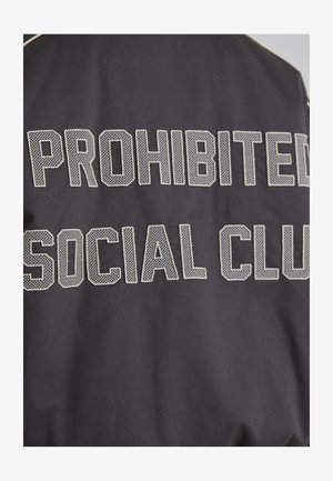 Jacheta neagră cu litere mari alb-negru, texturate cu plasă, care citește „SOCIAL CLUB PROHIBIT” pe spate. Material neted cu un finisaj mat.