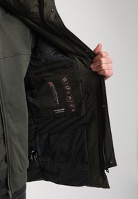 Interno della giacca verde oliva presenta una texture trapuntata, più tasche e simboli di cura stampati. L'esterno è liscio con una chiusura a zip.