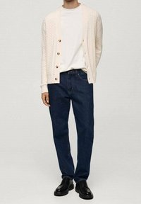 Beige cable-knit cardigan with wooden buttons over a white t-shirt, paired with dark blue jeans and black leather shoes.