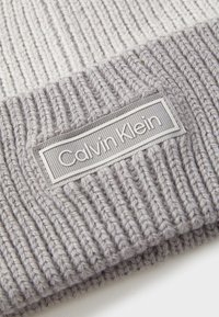 Grå strikket lue med ribbestruktur, med en rektangulær grå etikett med "Calvin Klein" i hvit skrift på forsiden.