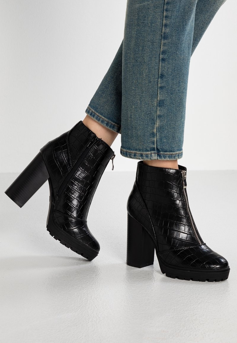 Bottes en cuir noir à texture croco, à talons hauts épais et zips avant, portées avec un jean en denim bleu.