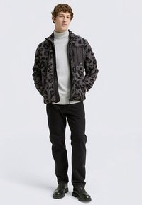 Fekete polárdzseki geometriai mintával, cipzáras záródással és mellzsebbel. Világosszürke garbó felett viselve, fekete farmerrel párosítva.