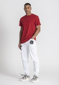 Röd bomullströja med broderad logga, tillsammans med vita slitna jeans med svarta accenter och mönstrade sneakers.